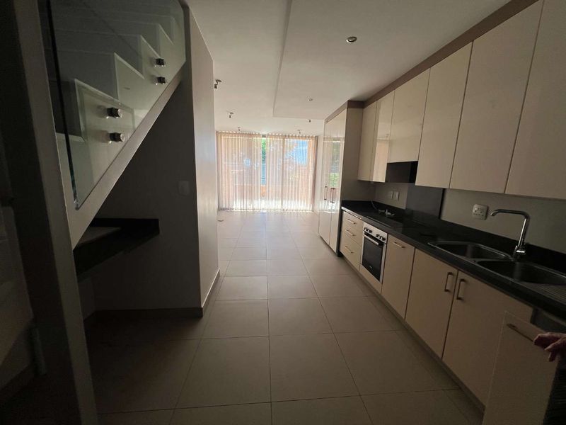 Modern open-plan kitchen featuring sleek cabinetry, integrated appliances, and tiled flooring, leading towards a bright living area with a glass-railed staircase.