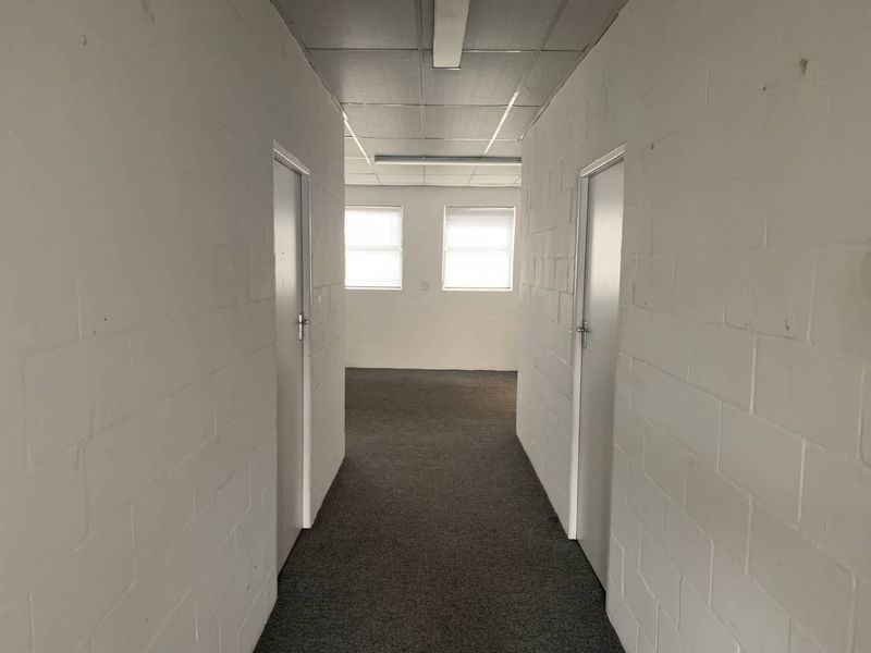 A functional hallway featuring painted brick walls, carpeted flooring, and a suspended ceiling with integrated lighting, leading into an open office or utility space.
