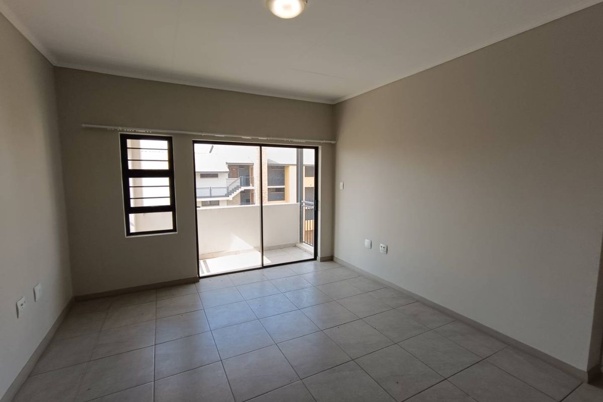 A bright, empty living area featuring neutral tiled flooring, a ceiling-mounted light fixture, and sliding glass doors leading to an outdoor balcony.
