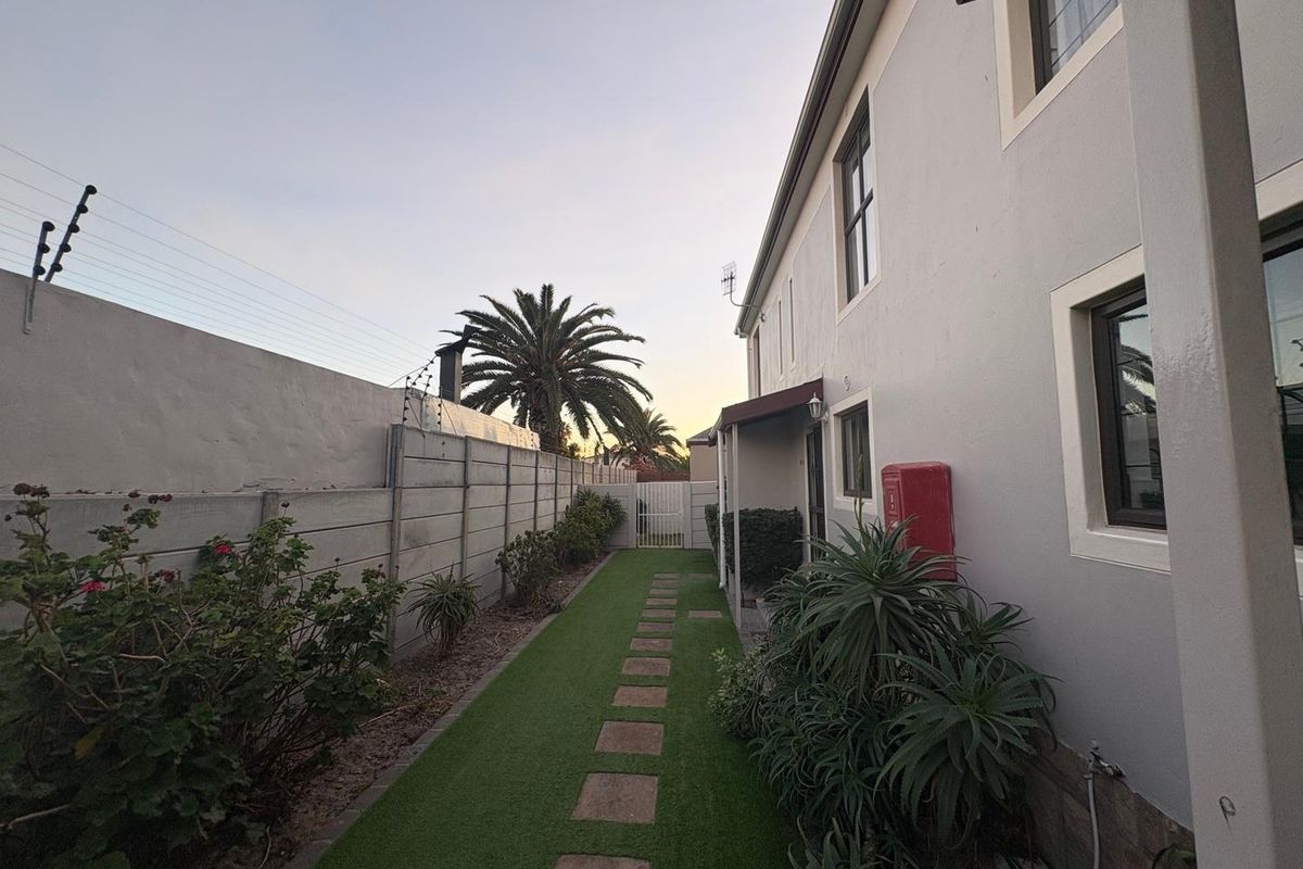 Side exterior view of the townhouse featuring a paved walkway, artificial lawn, perimeter wall with electric fencing, and established garden beds.