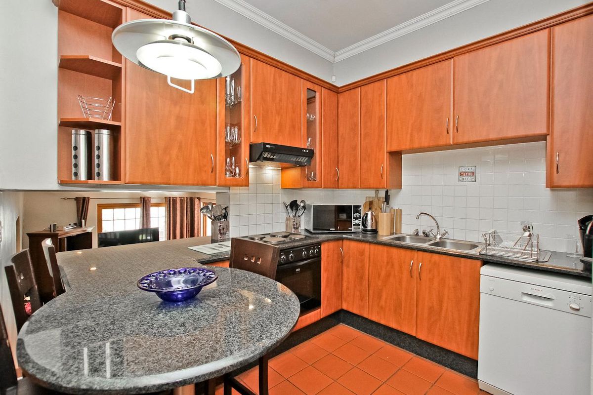 A functional kitchen featuring warm-toned wooden cabinetry, granite countertops, a built-in oven with hob, and a breakfast bar area with terracotta flooring.