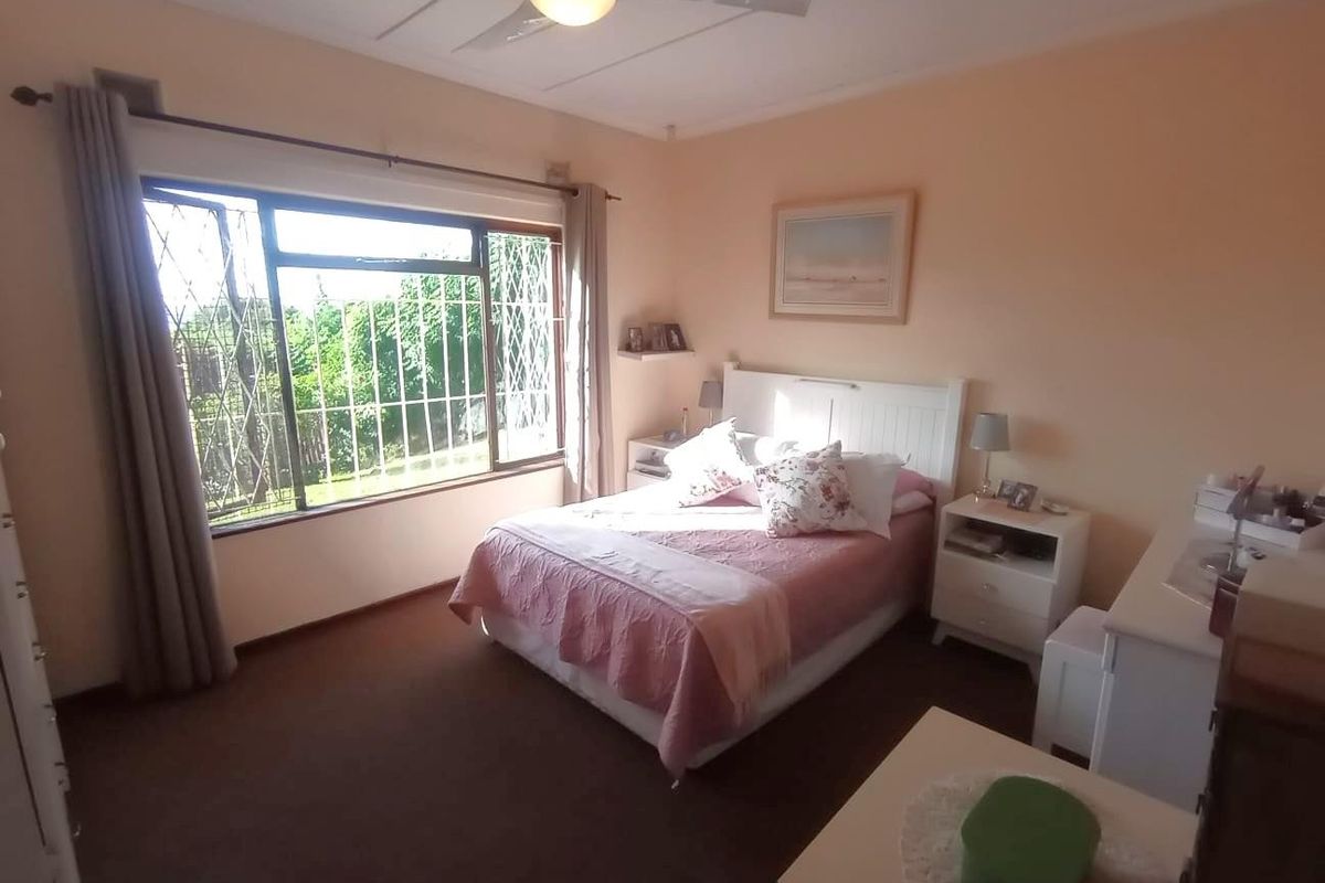 A standard bedroom featuring a ceiling fan, large window with security bars, and carpeted flooring.
