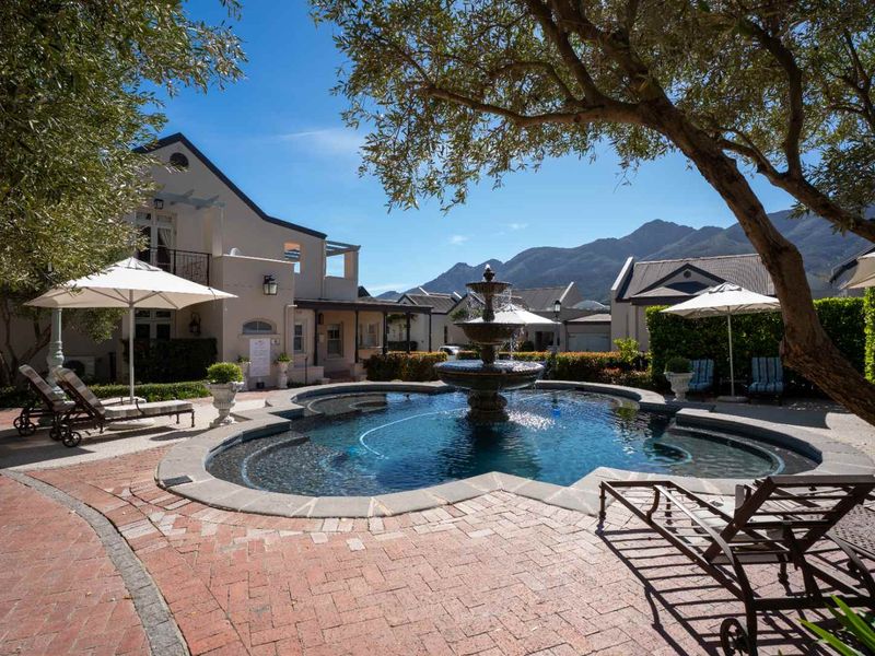 A beautifully landscaped pool area featuring a central stone fountain, brick paved surround, and mature trees set against a mountain backdrop.