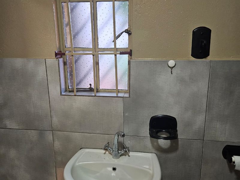 A functional bathroom featuring a pedestal basin with a chrome mixer tap, tiled walls, and a window fitted with security bars.