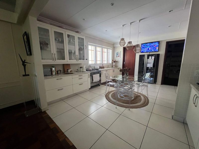 A bright, well-maintained kitchen featuring white cabinetry, glass-fronted display units, tiled flooring, and a dedicated dining area with modern pendant lighting.