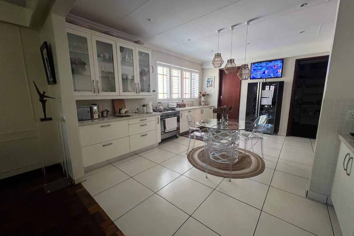 A bright, well-maintained kitchen featuring white cabinetry, glass-fronted display units, tiled flooring, and a dedicated dining area with modern pendant lighting.