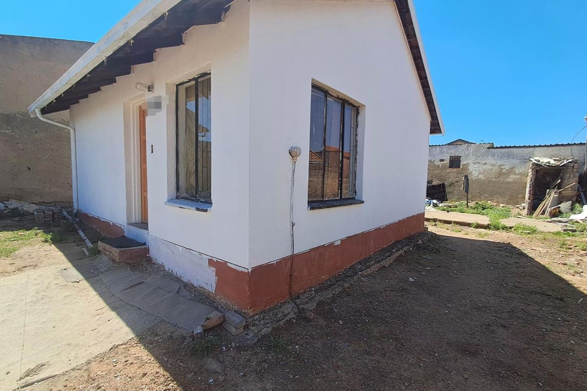 Rear view of a small detached dwelling featuring white painted walls, security bars on windows, and an unfinished exterior base.