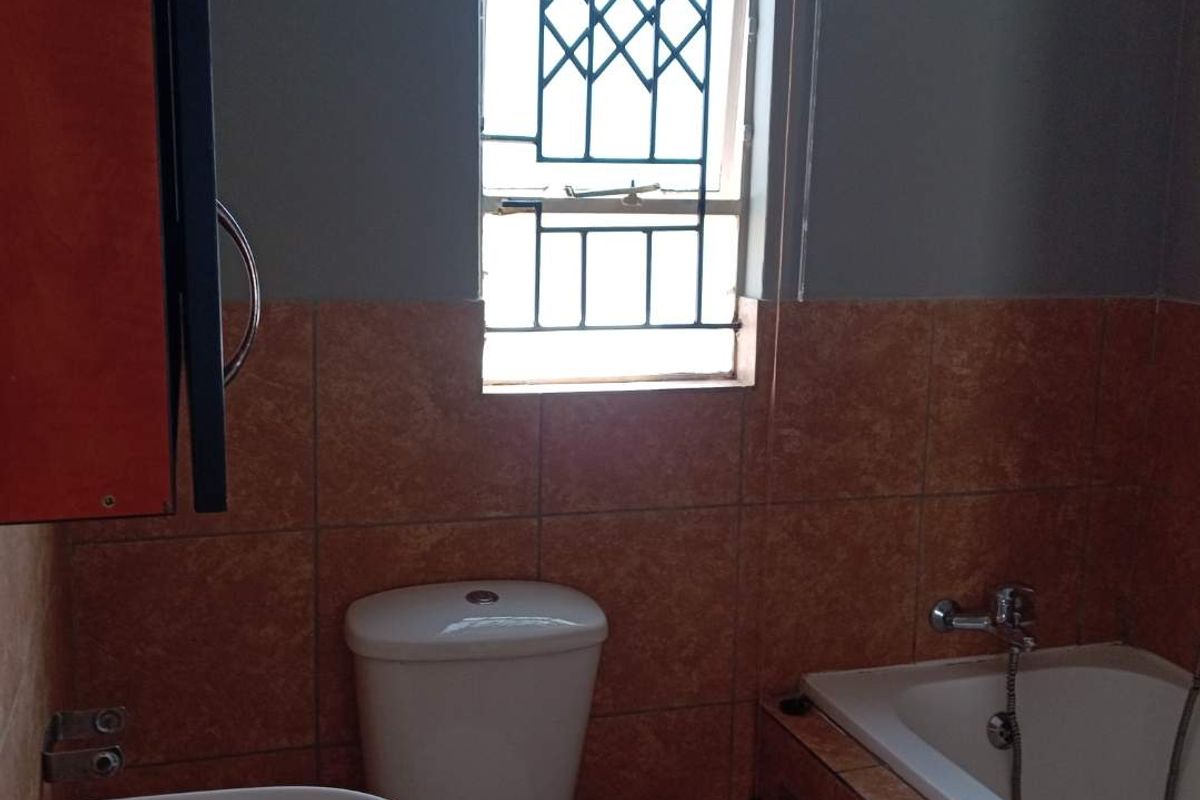 A functional bathroom featuring a bathtub, pedestal basin, and toilet, finished with mid-tone wall tiles and a security-grilled window.