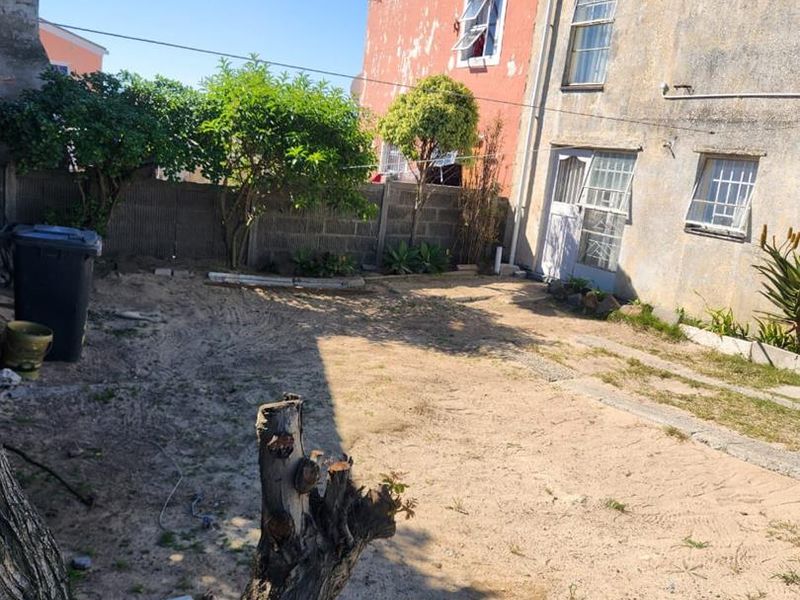 Rear exterior view of the property featuring a large sandy yard, boundary wall, and building facade showing signs of significant wear and peeling paint.