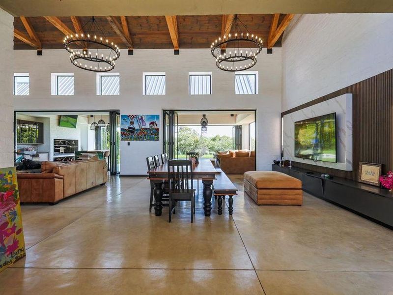 Spacious open-plan living and dining area featuring high vaulted ceilings with exposed timber beams, polished concrete floors, and seamless indoor-outdoor flow via bi-fold doors.