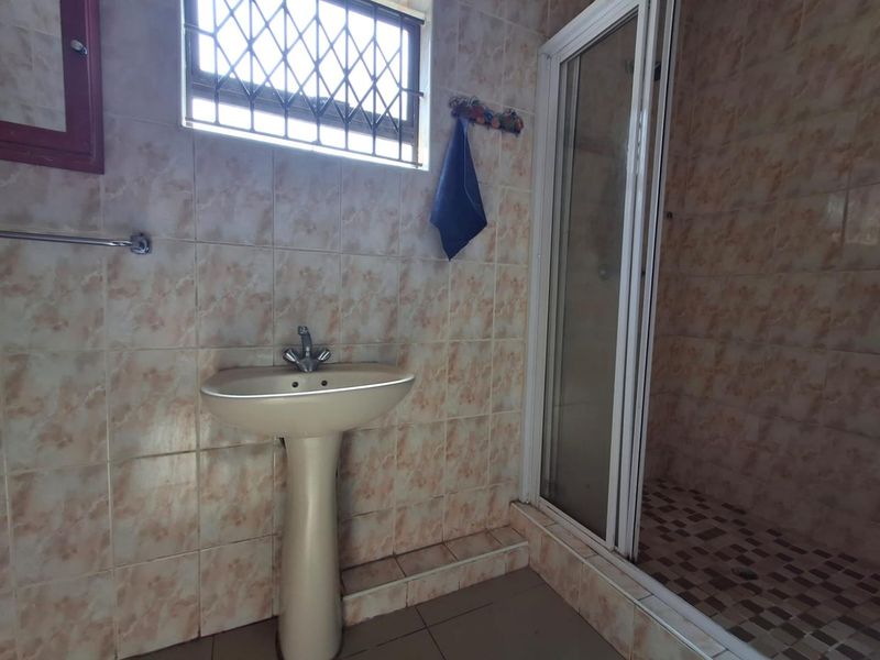 A functional bathroom featuring a pedestal basin, a shower enclosure, and dated wall tiling with a security-gated window.
