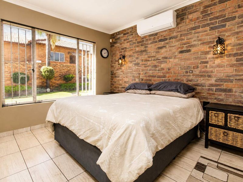 A well-maintained bedroom featuring an exposed brick accent wall, tiled flooring, and a split-system air conditioner, with large windows overlooking the garden.
