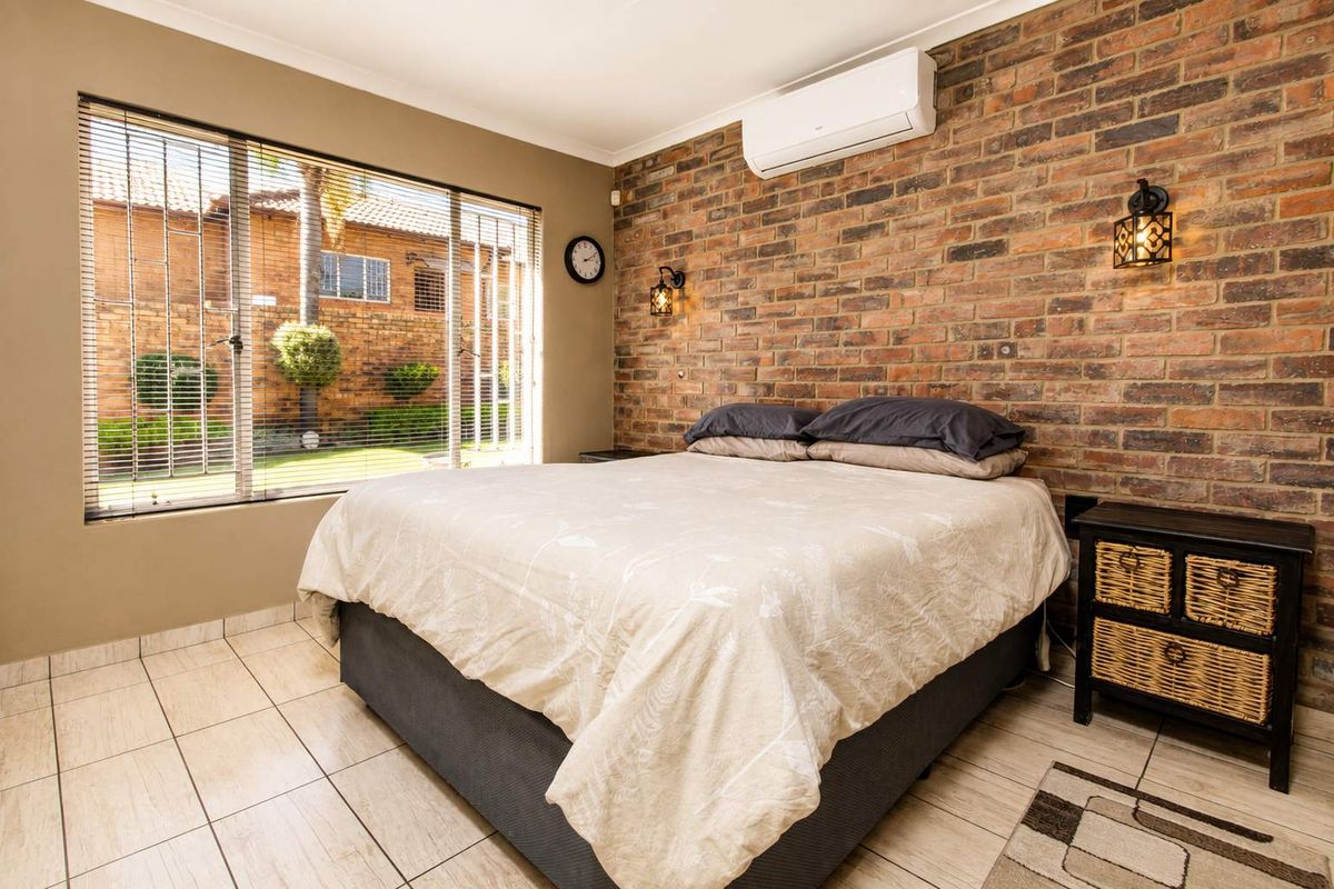 A well-maintained bedroom featuring an exposed brick accent wall, tiled flooring, and a split-system air conditioner, with large windows overlooking the garden.