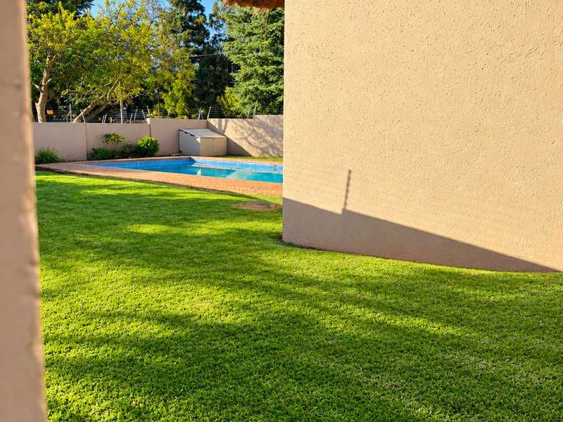 A well-maintained private garden featuring a swimming pool, lush green lawn, and a building with a traditional thatched roof.