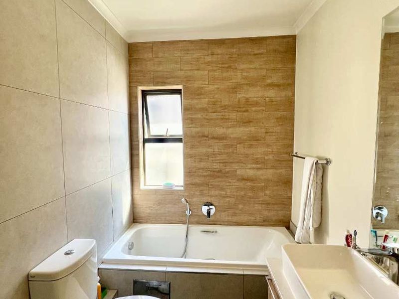 Modern bathroom featuring a built-in bathtub, wall-mounted basin with vanity cabinet, and a toilet, finished with neutral and wood-look wall tiling.