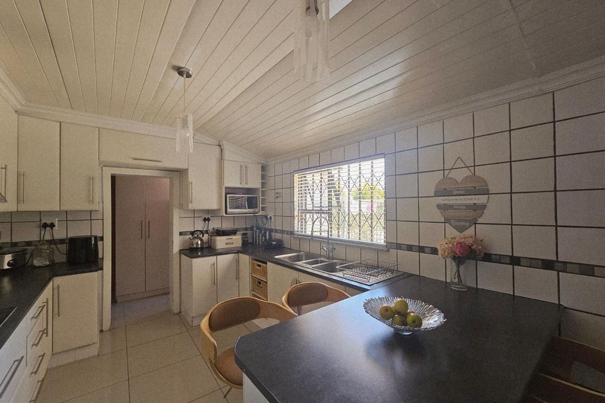 A functional kitchen featuring white cabinetry, dark countertops, tiled walls, and a unique tongue-and-groove ceiling, with an adjacent scullery area.