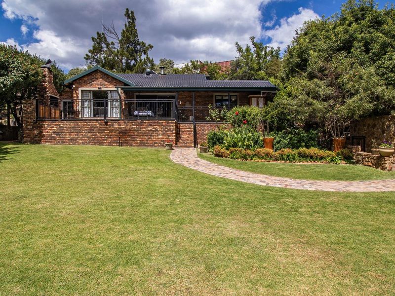 A well-maintained rear exterior view featuring a brick facade, raised patio with metal railings, built-in outdoor fireplace, and a landscaped garden with paved walkway.