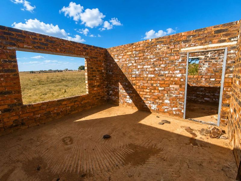 An unfinished room structure featuring exposed brick walls, a metal door frame, and open window spaces, set within a rural farm landscape.
