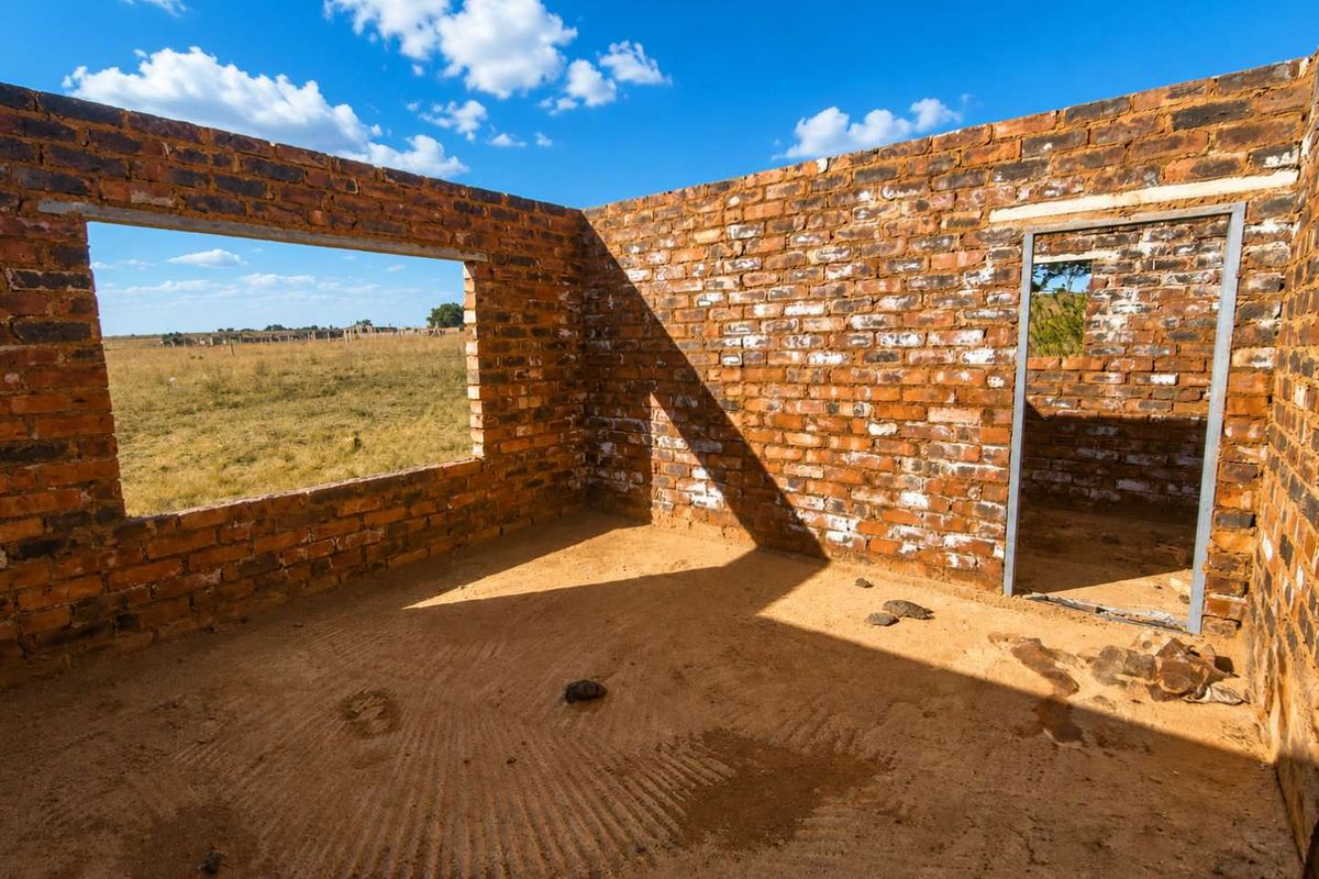 An unfinished room structure featuring exposed brick walls, a metal door frame, and open window spaces, set within a rural farm landscape.