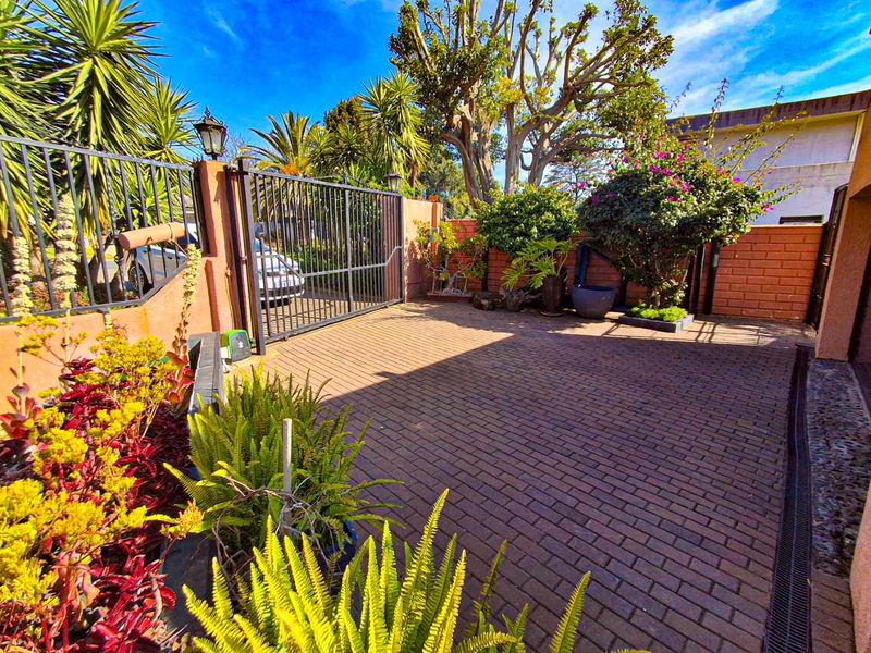 A well-maintained paved driveway entrance featuring a secure metal gate, brick boundary walls, and established garden landscaping.
