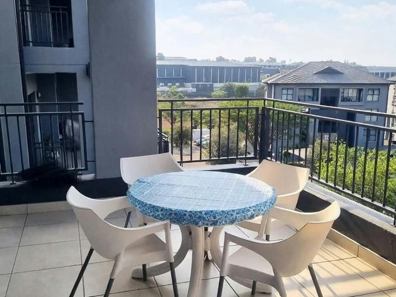 A spacious covered balcony featuring tiled flooring and a metal balustrade, offering an open view of the surrounding residential complex.