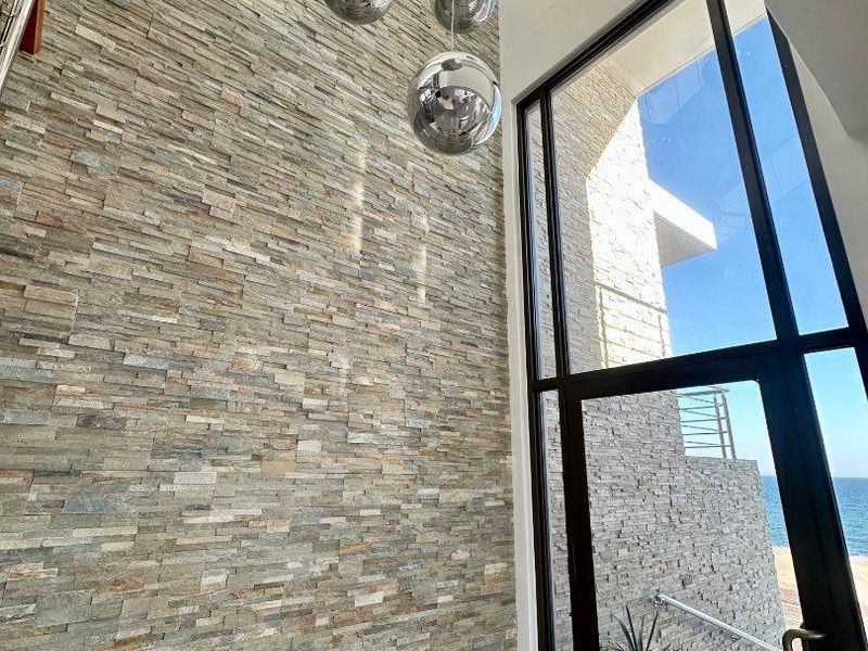 A grand entrance hall featuring a striking floor-to-ceiling stacked stone feature wall, expansive glass windows, and modern pendant lighting with ocean views.