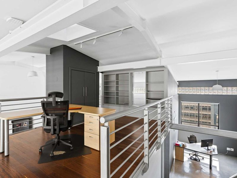 A modern mezzanine office space featuring exposed structural beams, hardwood flooring, built-in shelving, and a sleek metal balustrade overlooking the lower level.