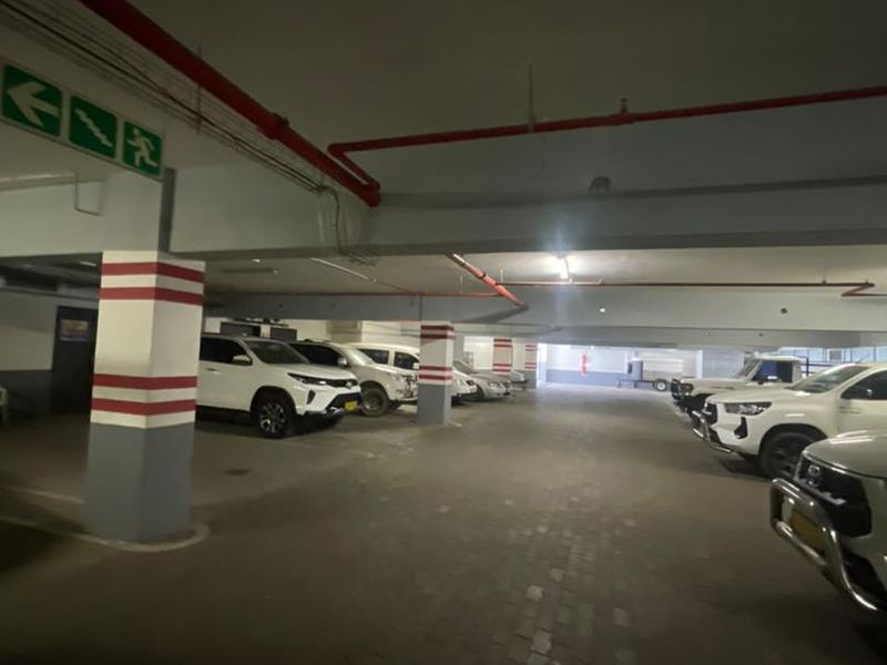 Undercover parking garage area featuring concrete structural columns, overhead fire suppression piping, and paved flooring.
