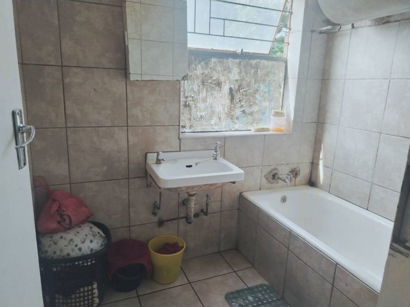 A dated bathroom featuring a wall-mounted basin and bathtub with tiled finishes, requiring significant renovation and repair to the window and wall surfaces.