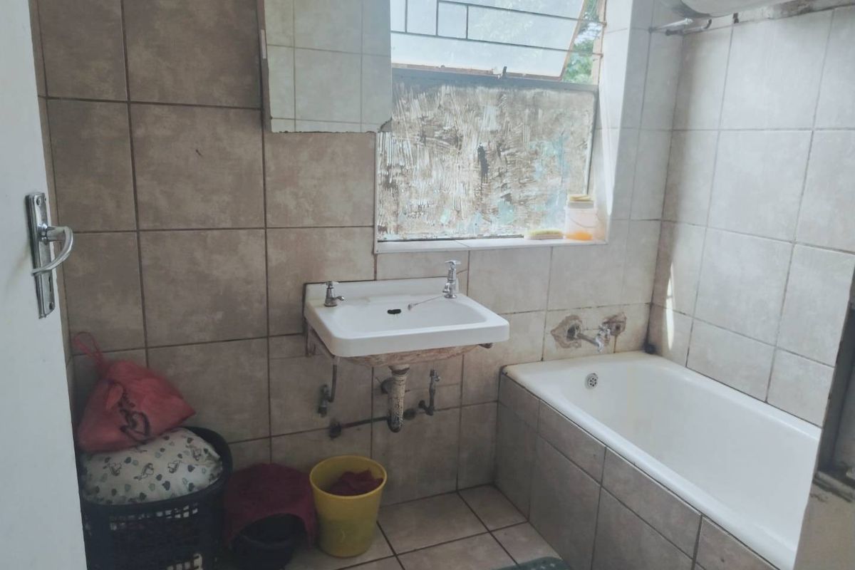 A dated bathroom featuring a wall-mounted basin and bathtub with tiled finishes, requiring significant renovation and repair to the window and wall surfaces.