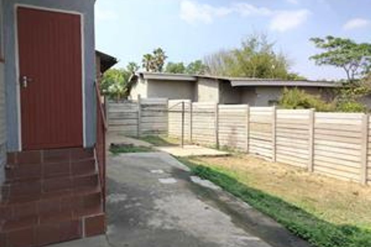 A rear exterior view of the property featuring a concrete patio area, a red entrance door with steps, and a perimeter precast concrete wall.
