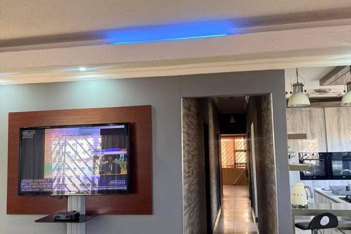 A modern living area featuring a built-in television wall unit, decorative ceiling lighting, and an open-plan transition into the kitchen and hallway.
