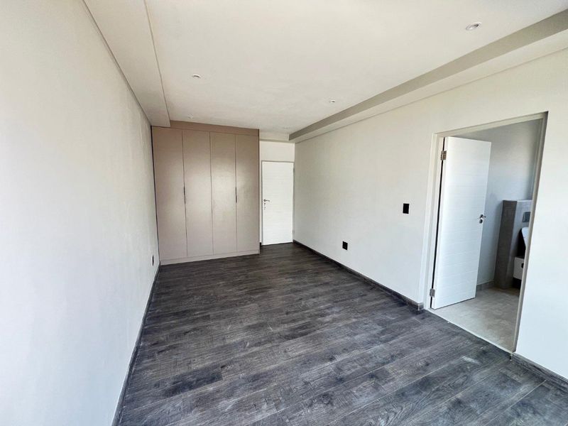 A bright, empty bedroom featuring dark wood-look flooring, built-in cupboards, and an adjoining en-suite bathroom.