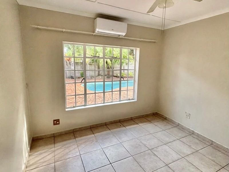An empty bedroom featuring tiled flooring, a ceiling fan, and a wall-mounted air conditioning unit, with a view overlooking the outdoor pool area.