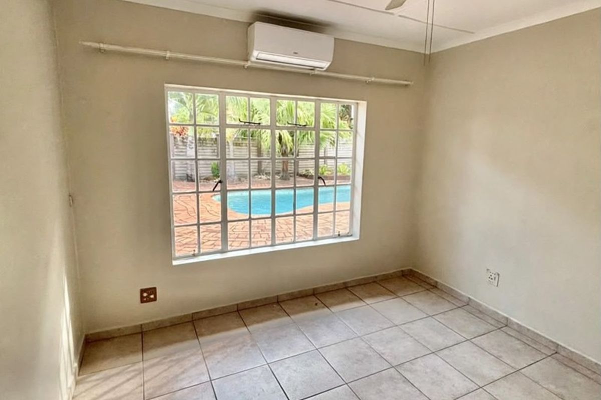 An empty bedroom featuring tiled flooring, a ceiling fan, and a wall-mounted air conditioning unit, with a view overlooking the outdoor pool area.