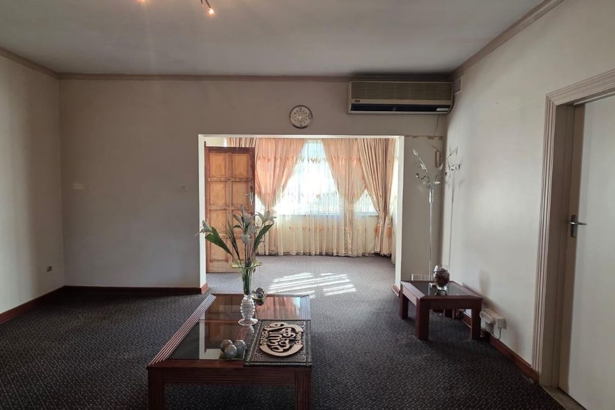 A spacious living room featuring carpeted flooring, a wall-mounted air conditioning unit, and an open transition to an adjacent sunlit area.