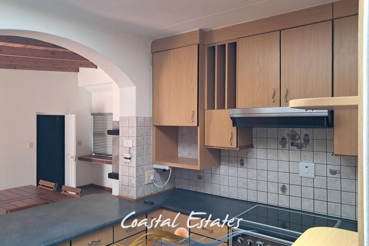 A functional kitchen featuring light wood-finish cabinetry, a built-in oven with hob, extractor fan, and tiled backsplash, connected to an adjacent living area via archway.