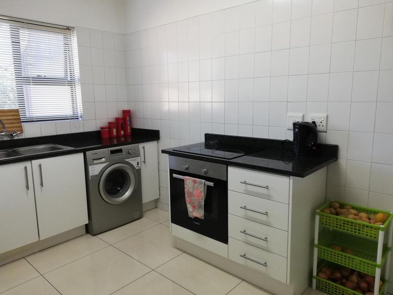 A functional kitchen featuring white cabinetry, granite countertops, a built-in oven with hob, and an integrated washing machine, finished with tiled splashbacks and flooring.