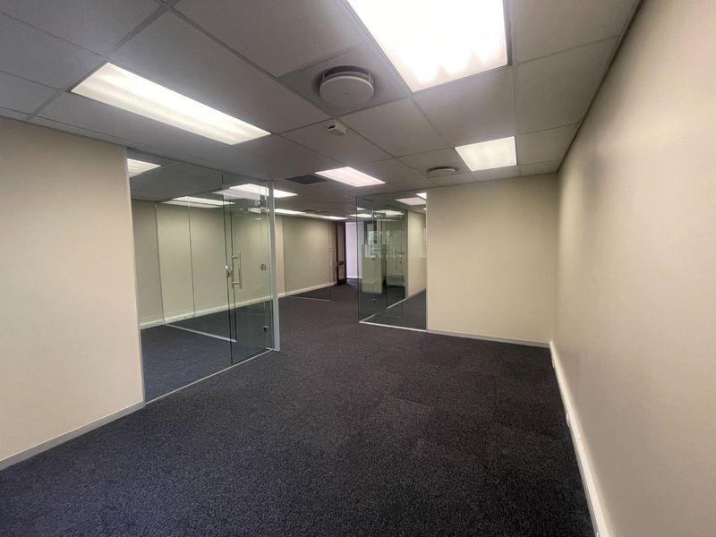 A vacant office space featuring glass partitions, carpeted flooring, and a suspended ceiling with recessed lighting, offering a clean and professional work environment.