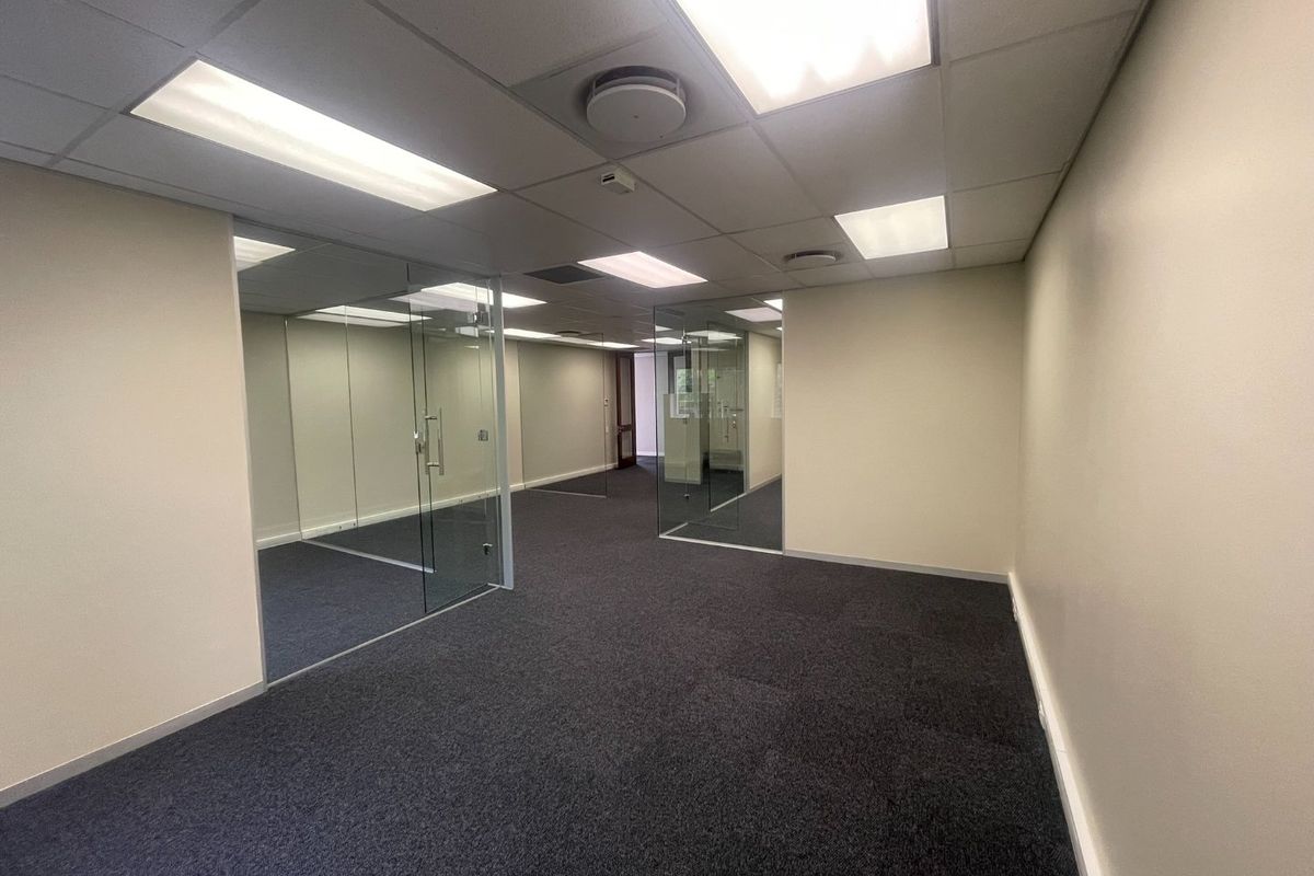 A vacant office space featuring glass partitions, carpeted flooring, and a suspended ceiling with recessed lighting, offering a clean and professional work environment.
