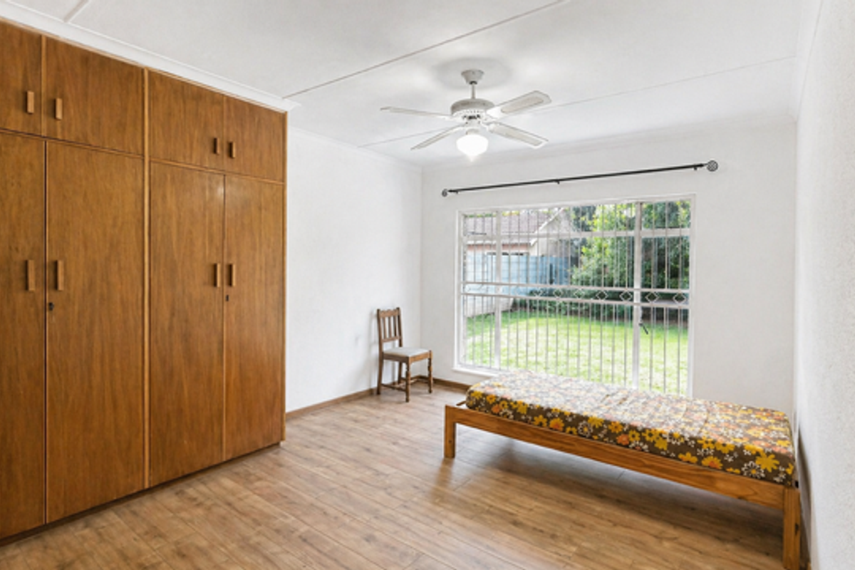 A bedroom featuring built-in wooden wardrobes, wood-look flooring, a ceiling fan, and a large window with security bars overlooking the garden.