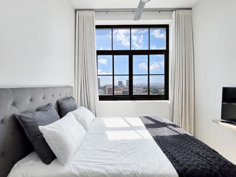 A bright, modern bedroom featuring a large industrial-style window with expansive city views and a ceiling fan.
