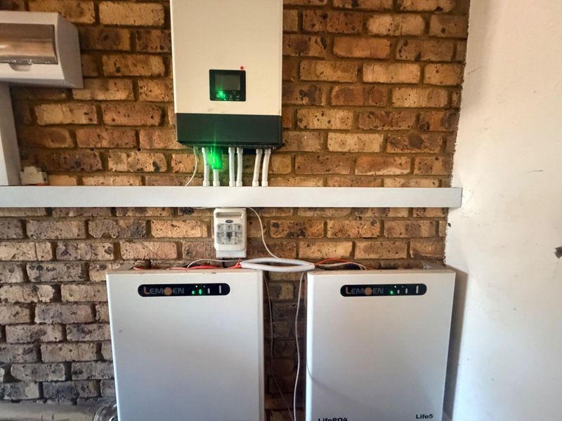 A utility area featuring a wall-mounted solar inverter and dual lithium-ion battery storage units installed against an exposed brick wall.