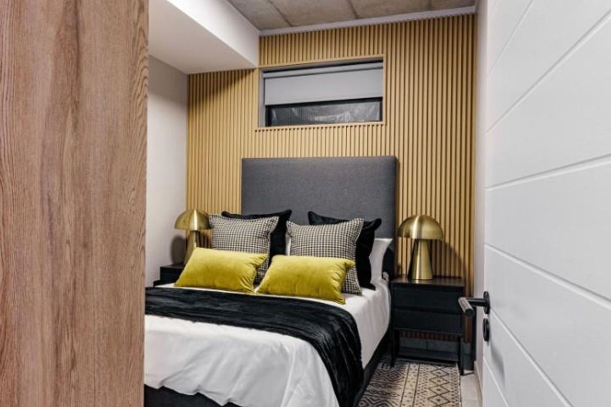 A modern bedroom featuring a decorative timber slat feature wall, exposed concrete ceiling, and a high-set window with a roller blind.