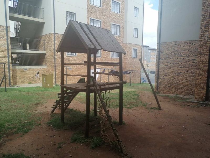 Communal outdoor area featuring a face brick townhouse complex facade, metal stairwells, and a small wooden playground structure set on a patchy lawn.