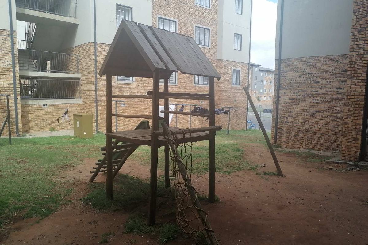 Communal outdoor area featuring a face brick townhouse complex facade, metal stairwells, and a small wooden playground structure set on a patchy lawn.