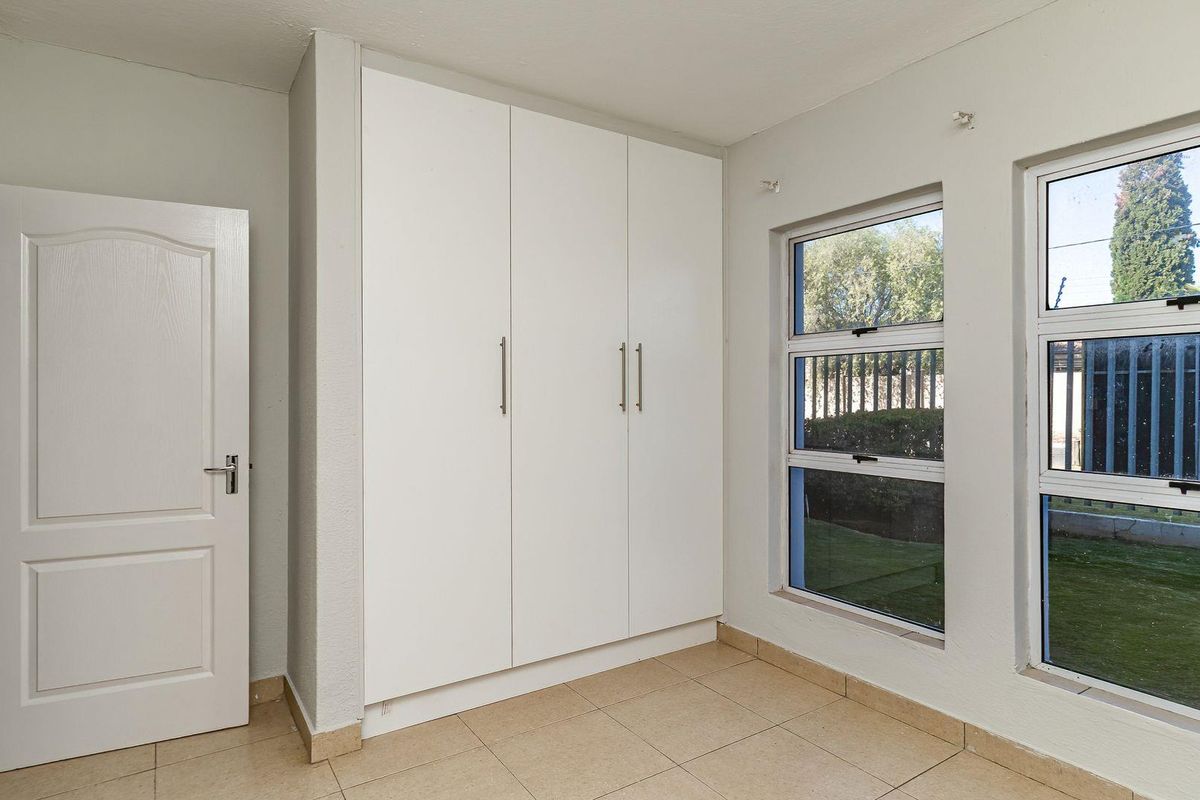 A bright, empty bedroom featuring neutral tiled flooring, built-in white cupboards, and large windows overlooking the garden area.