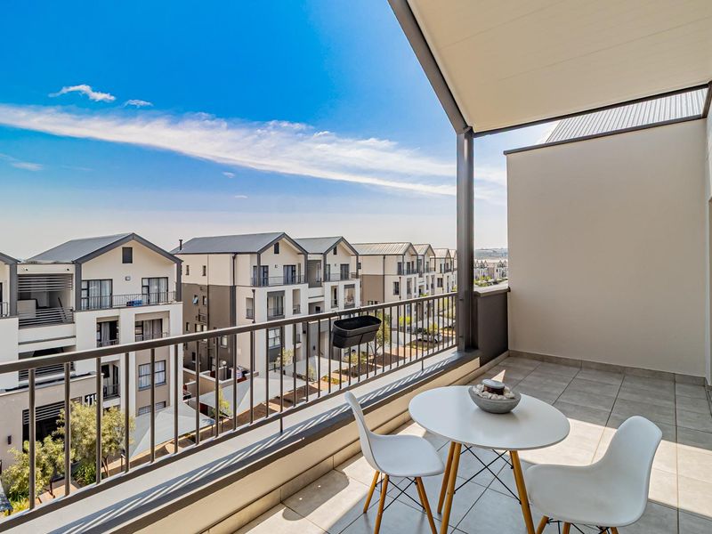 A spacious covered balcony featuring tiled flooring and a metal balustrade, offering an elevated view of the surrounding residential complex.