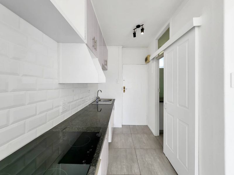 A clean, modern galley kitchen featuring a subway tile backsplash, granite countertops, built-in electric hob, and neutral tiled flooring.
