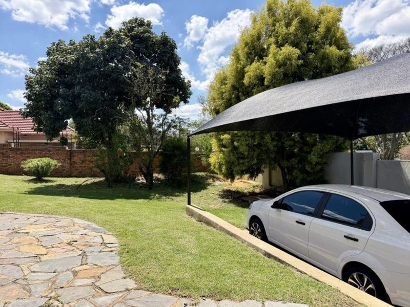 A rear garden area featuring a stone paved path, a lawn, and a covered parking space with a shade sail structure.